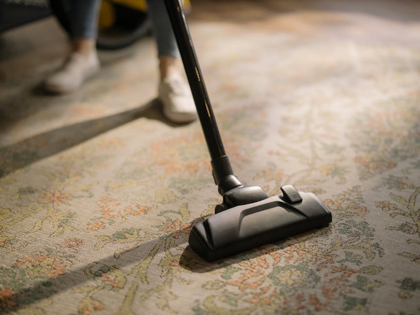 A woman with curly hair wearing a beige jacket, dark jeans, and white shoes is operating a yellow vacuum cleaner with a black hose and a wide cleaning nozzle on a patterned area rug in a living room.